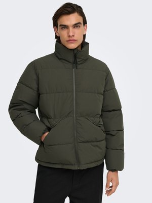 Jeune homme portant une veste matelassée vert foncé avec un col haut et les mains dans les poches avant, debout devant un fond clair uni.