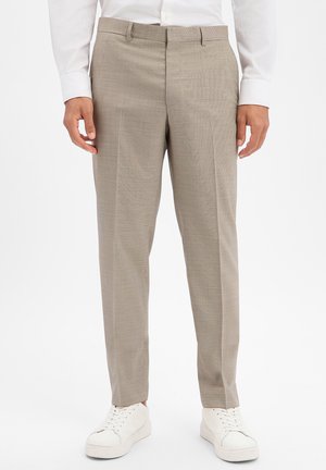 Mann trägt beige maßgeschneiderte Hose mit vorderen Bügelfalten, kombiniert mit weißen Sneakers und einem weißen Langarmhemd, steht vor weißem Hintergrund.