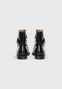 Botas de tornozelo de verniz preto com um acabamento liso, apresentando um detalhe de fivela dourada e um salto baixo. Fecho com zíper na parte traseira para fácil acesso.
