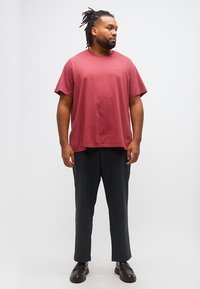 Άνδρας με ντραντλοκς που φοράει κόκκινο κοντομάνικο μπλουζάκι, μαύρο παντελόνι και σκούρα μοκασίνια, στέκεται και κοιτάζει προς τα δεξιά του.