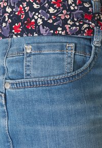 Ljusblå denimjeans med en liten framficka och metallknapp, med ett midjeband i mörkt blommönster. Slät textur.