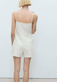 Hellbeige Jumpsuit mit dünnen Trägern, quadratischem Rücken, kurzen Beinen und glatter Baumwolltextur. Einfaches, minimalistisches Design.