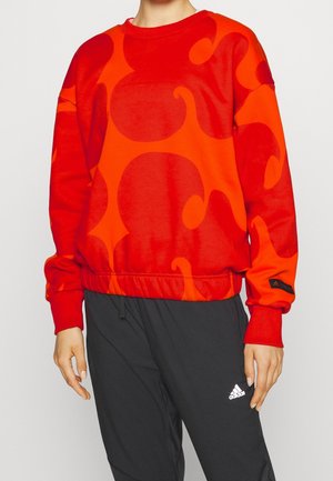 Osoba nosząca czerwoną i pomarańczową abstrakcyjną wzorzystą bluzę z długim rękawem oraz czarne spodnie Adidas z białym logo.