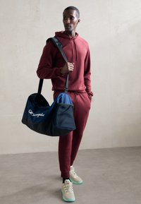 Rød hettegenser og joggebukser, holder en blå og svart treningsbag med hvit merkevare, parret med beige joggesko med teal såler.