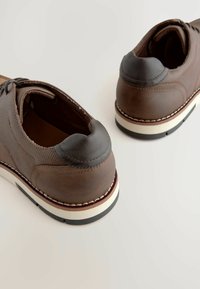 Zapatos de cuero marrón con un panel lateral texturizado, puntera redondeada y suela de goma blanca contrastante con ribete negro. Detalles suaves en el tacón.