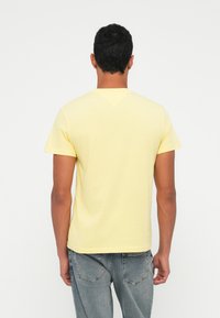 Geltonas medvilninis marškinėliai trumpomis rankovėmis ir V formos iškirpte, matomi iš nugaros. Sukomplektuoti su šviesiai pilkais džinsais, pabrėžiančiais laisvą stilių.