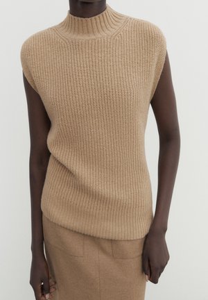 Personne portant un pull sans manches beige en tricot avec un col montant et une jupe beige assortie sur un fond uni.
