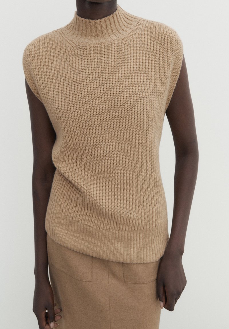 Personne portant un pull sans manches beige en tricot avec un col montant et une jupe beige assortie sur un fond uni.