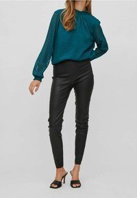 Blusa a maniche lunghe color teal con motivo a zigzag testurizzato, abbinata a pantaloni in pelle nera aderenti e tacchi aperti neri.