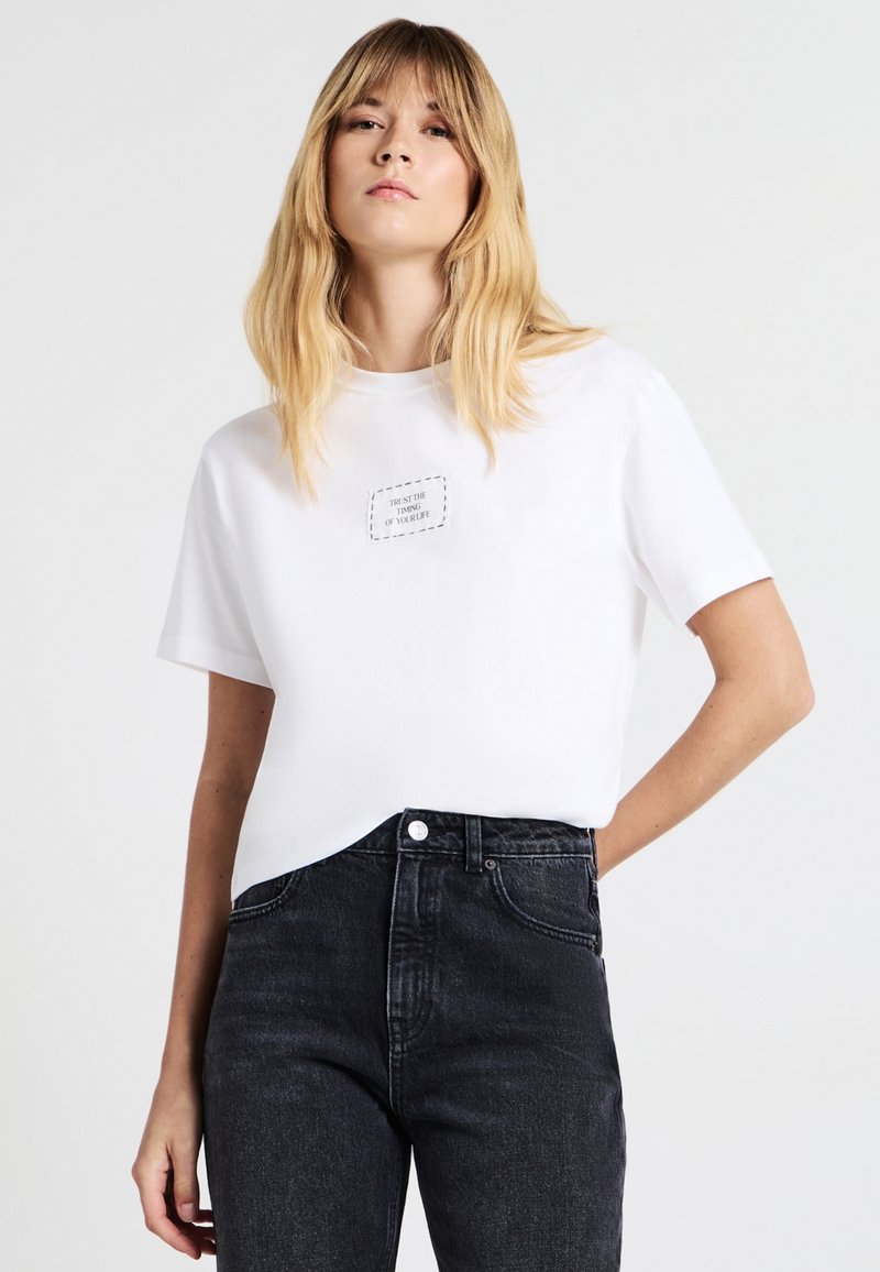 T-shirt bianco in cotone con maniche corte, con un'etichetta rettangolare cucita con testo sul davanti. Abbinato a jeans neri a vita alta.