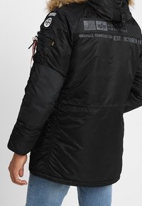 Veste isolante noire avec capuche bordée de fourrure, équipée de poignets élastiques, de patchs et de texte graphique au dos. Tissu en nylon texturé.