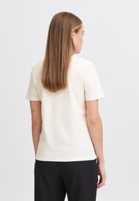 T-shirt en coton crème clair avec des manches courtes et un col rond, présentant une coupe décontractée. Porté avec un pantalon noir, sans motifs ni accents visibles.