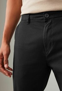 Pantalons noirs en tissu texturé avec une coupe slim, dotés d'une fermeture à bouton et de passants de ceinture, présentés au niveau de la taille et des hanches.