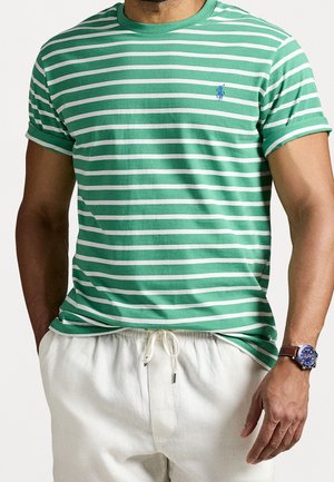 Mężczyzna w zielono-białej koszulce w paski z niebieskim logo, białych spodniach na sznurku oraz zegarku na rękę z niebieską tarczą i brązowym paskiem.