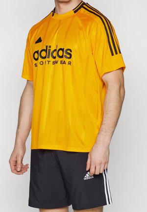 Mężczyzna w żółtej sportowej koszulce Adidas z czarnymi paskami i czarnych szortach Adidas z białymi paskami, stojący na jednolitym tle.