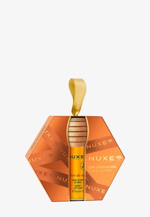 Sześciokątne pomarańczowe pudełko z złotą wstążką, zawierające żółtą tubkę balsamu do ust z drewnianym korkiem, oznaczoną napisem "Pielęgnacja warg z miodem".