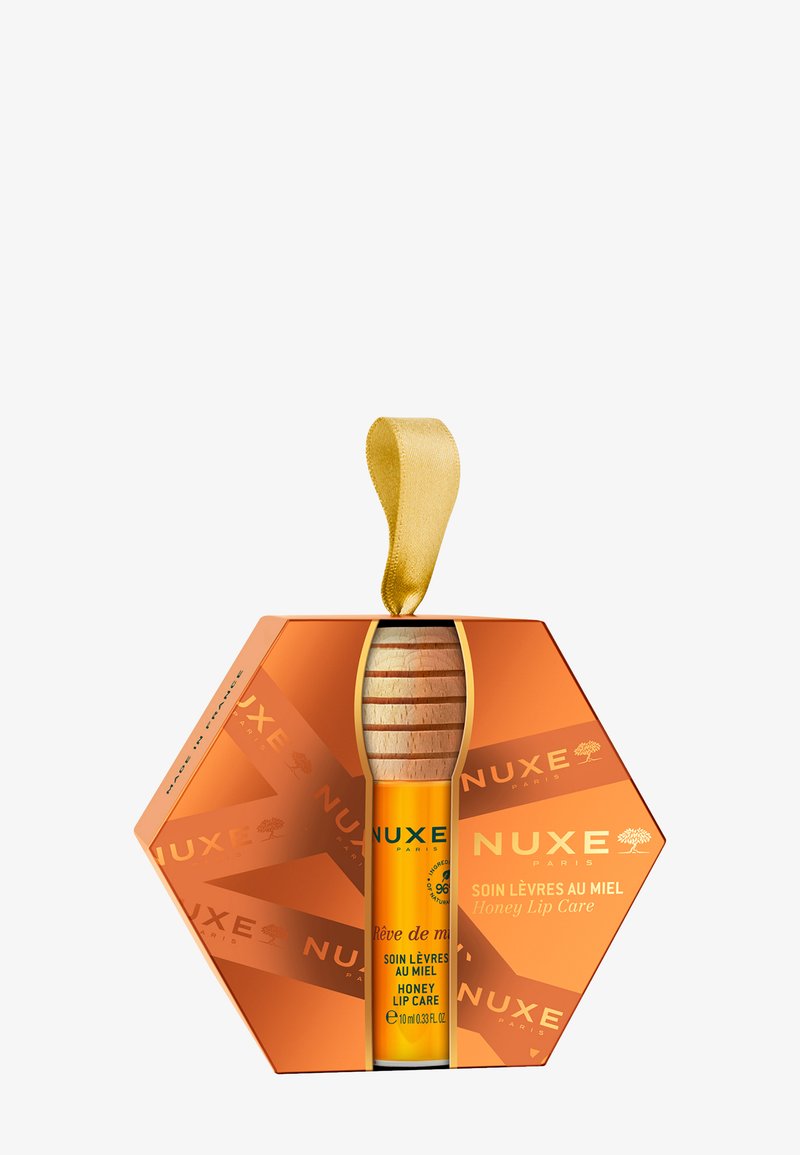 Sześciokątne pomarańczowe pudełko z złotą wstążką, zawierające żółtą tubkę balsamu do ust z drewnianym korkiem, oznaczoną napisem "Pielęgnacja warg z miodem".