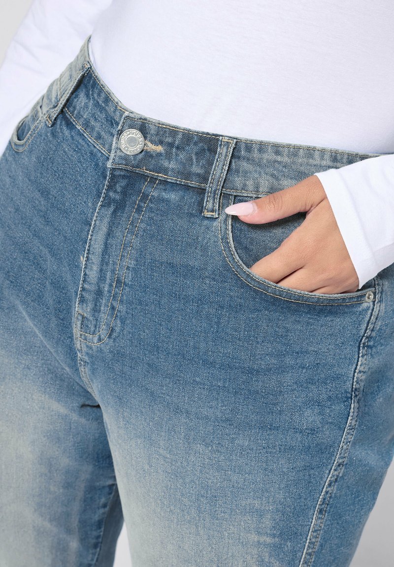 Ljusblå denimjeans med blekningar, fem fickor, silverknappdetalj och synlig söm längs sömmarna. Hand i fickan.