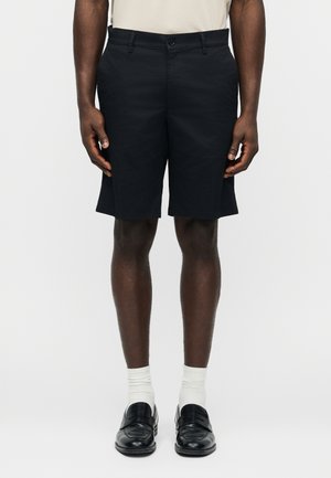 Mann trägt schwarze maßgeschneiderte Shorts, weiße Socken und schwarze Lederloafers, steht vor einem schlichten hellen Hintergrund.