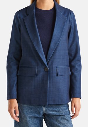 Persoon die een blauw geruit colbert draagt met inkepingkraag, een enkele zwarte knoop, klepzakken, een navy trui en blauwe spijkerbroek.