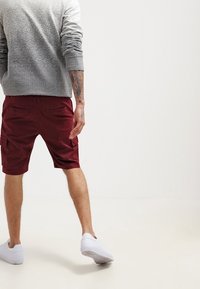 Sweater gris texturé, shorts bordeaux avec poches latérales et baskets blanches. Les shorts sont de longueur genou avec une coupe décontractée.