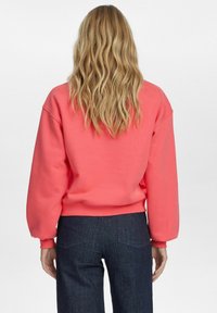 Korall színű pulóver laza szabással, bordás mandzsettákkal és aljjal. Sötét farmernadrággal párosítva, ami egy laza, kényelmes stílust mutat.