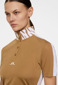 Brązowa koszulka polo z wysokim kołnierzem, w biało-brązowy wzór zebry, z zapięciem na guziki z przodu oraz białymi akcentami po bokach.