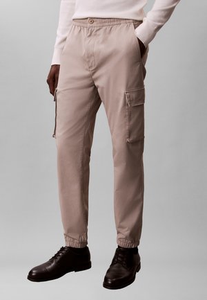 Hombre lleva pantalones cargo beige con puños elásticos, camiseta blanca de manga larga y zapatos de vestir de cuero negro, de pie frente a un fondo liso.