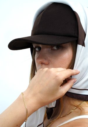 Brown felt cap with a white scarf featuring brown accents. Hand positioned near face, wearing a gold chain bracelet on wrist.