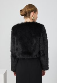 Kvinne med hår i en knute, iført store gulløredobber og en svart faux pelsjakke, fotografert bakfra mot en hvit bakgrunn.