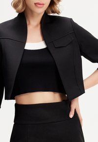 Veste noire courte avec un col haut et une poche poitrine unique, associée à un crop top noir à côtes avec un col blanc.