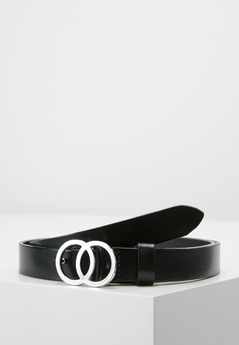 Ceinture en cuir noir avec boucle double anneau argentée, enroulée et posée sur un piédestal blanc sur un fond uni.