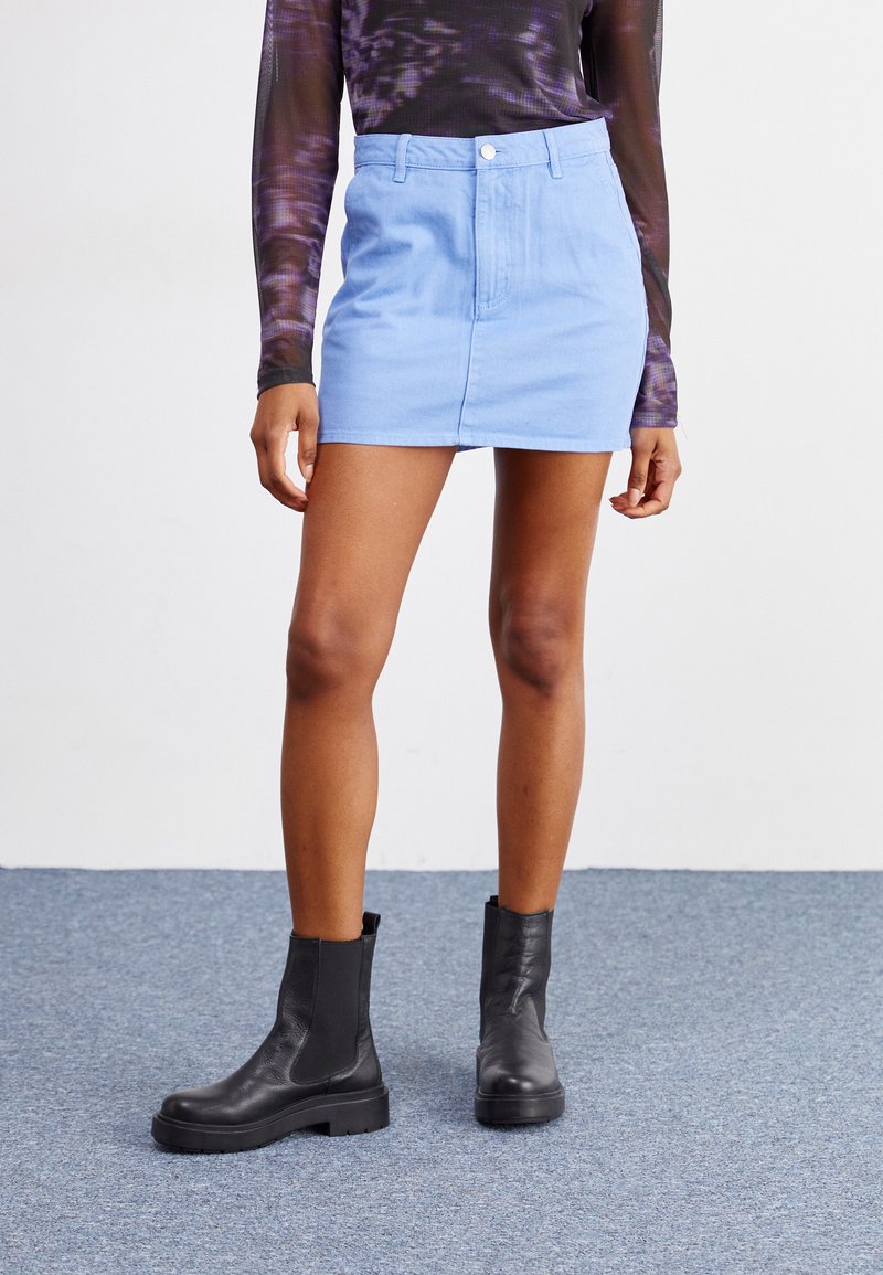Jupe en denim bleu courte avec une coupe droite, poche avant et fermeture à boutons. Assortie à des bottes à talons épais noires.