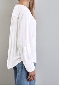 Chemise blanche à manches longues avec un dos plissé, des poignets boutonnés et un ourlet arrondi, associée à un denim bleu clair. Tissu lisse et léger.