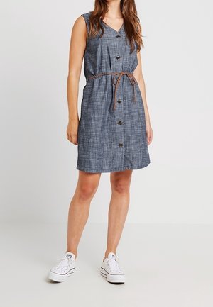 Femme portant une robe bleue boutonnée sans manches, longueur genou, avec une fine ceinture marron et des baskets blanches, debout sur un fond uni.