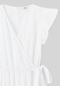 Robe blanche avec des manches courtes à volants, présentant un design croisé, une taille élastique et un motif à œillets. Liens sur le côté gauche, tissu texturé.