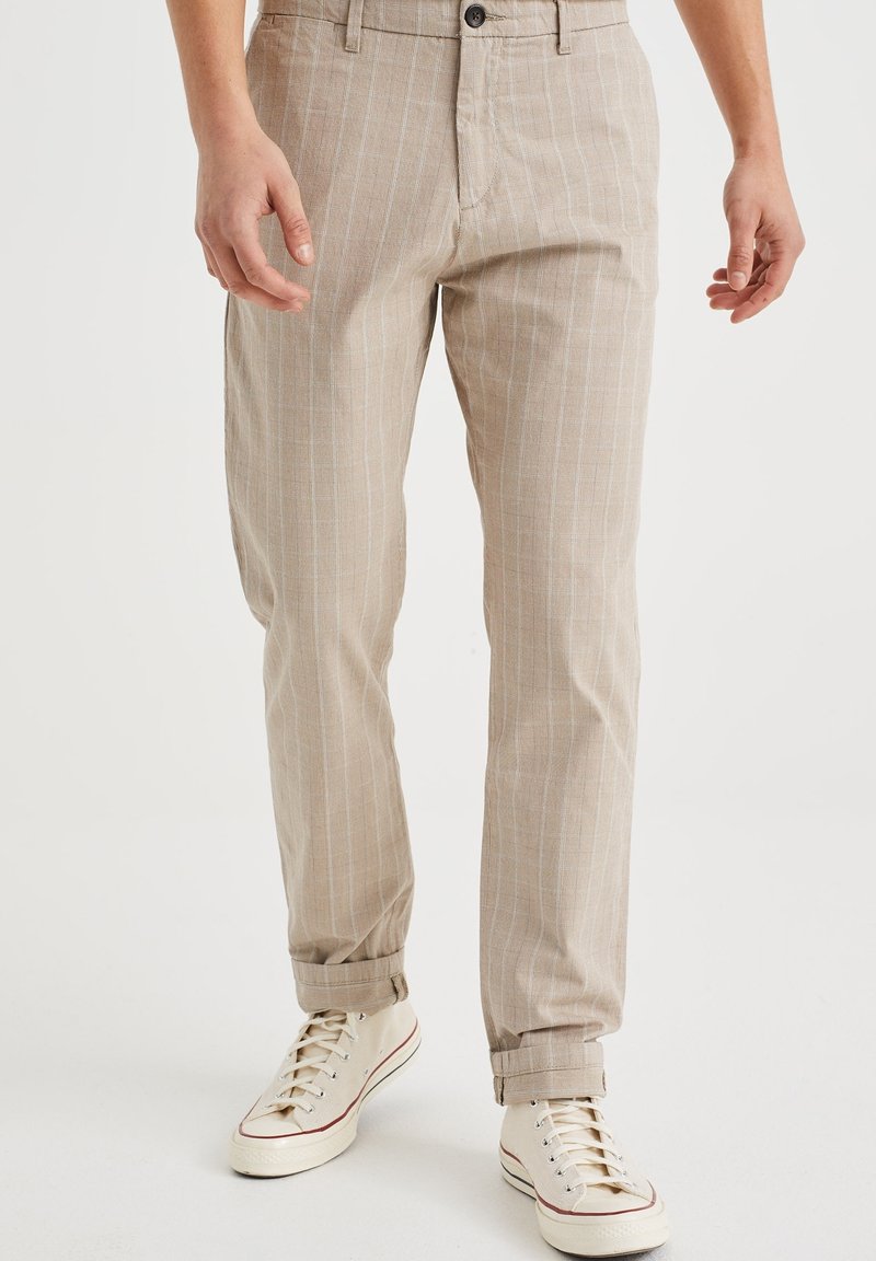 Pantalons en lin beige avec des rayures bleu clair, une fermeture à bouton et des ourlets roulés, associés à des baskets en toile blanches.