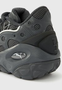 Κοντινή λήψη της φτέρνας ενός μαύρου γκρι αθλητικού παπουτσιού Puma με υφή στη σόλα, επάνω μέρος από δίχτυ, κυκλικές λεπτομέρειες ραφής και ορατό λογότυπο στη μεσαία σόλα.