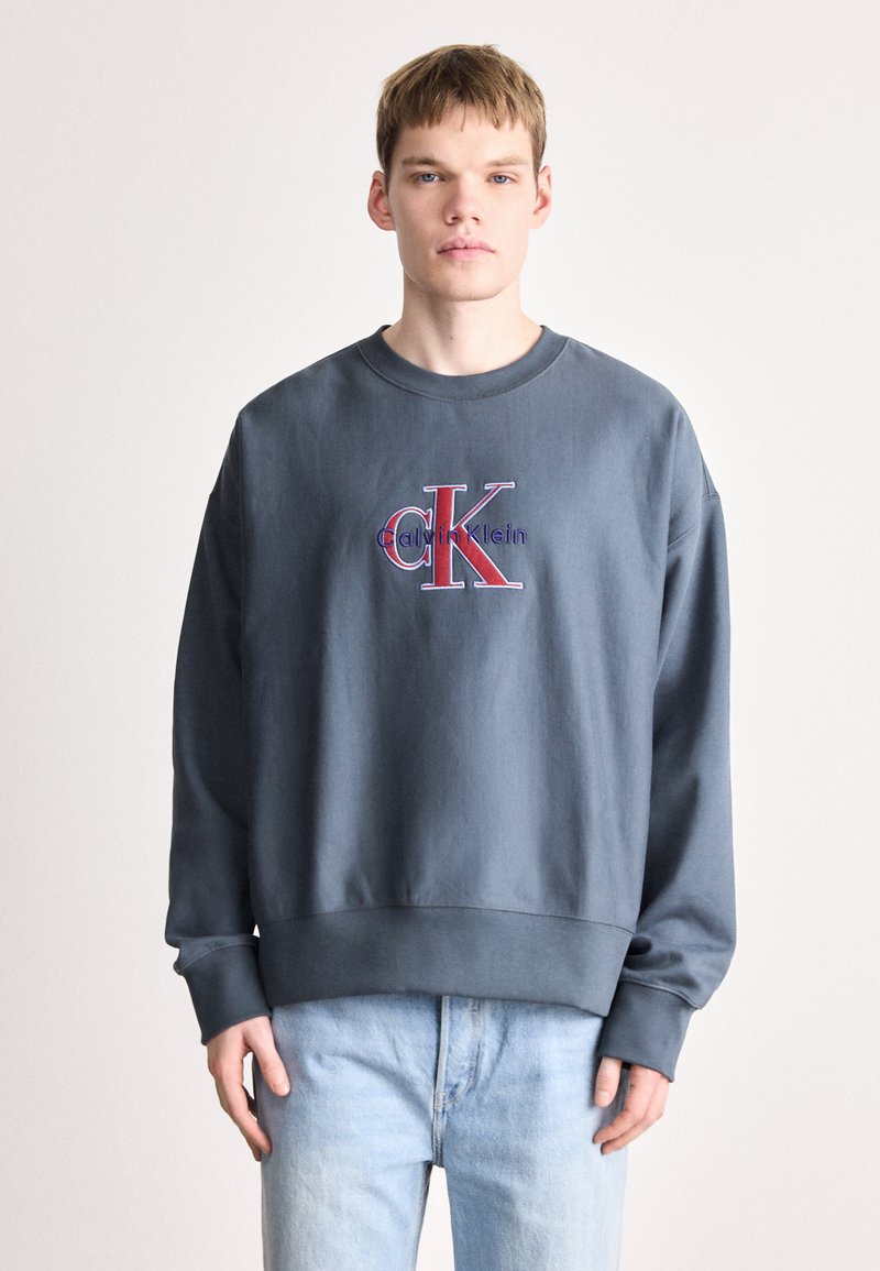 Jovem vestindo um moletom cinza escuro da Calvin Klein com um logótipo vermelho e azul, combinado com jeans azul claro, posando frente a um fundo simples.