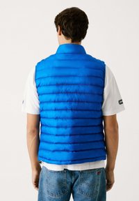 Polstret blå vest med høj krave, udstyret med vertikale quiltede sektioner. Bæret over en hvid t-shirt og parret med blå jeans.