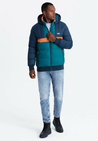 Praktisk jacka med vadderad design i teal, mörkblå ärmar och ett orange horisontellt rand. Inkluderar huva och dragkedja. Bärs med ljusa jeans och stövlar.