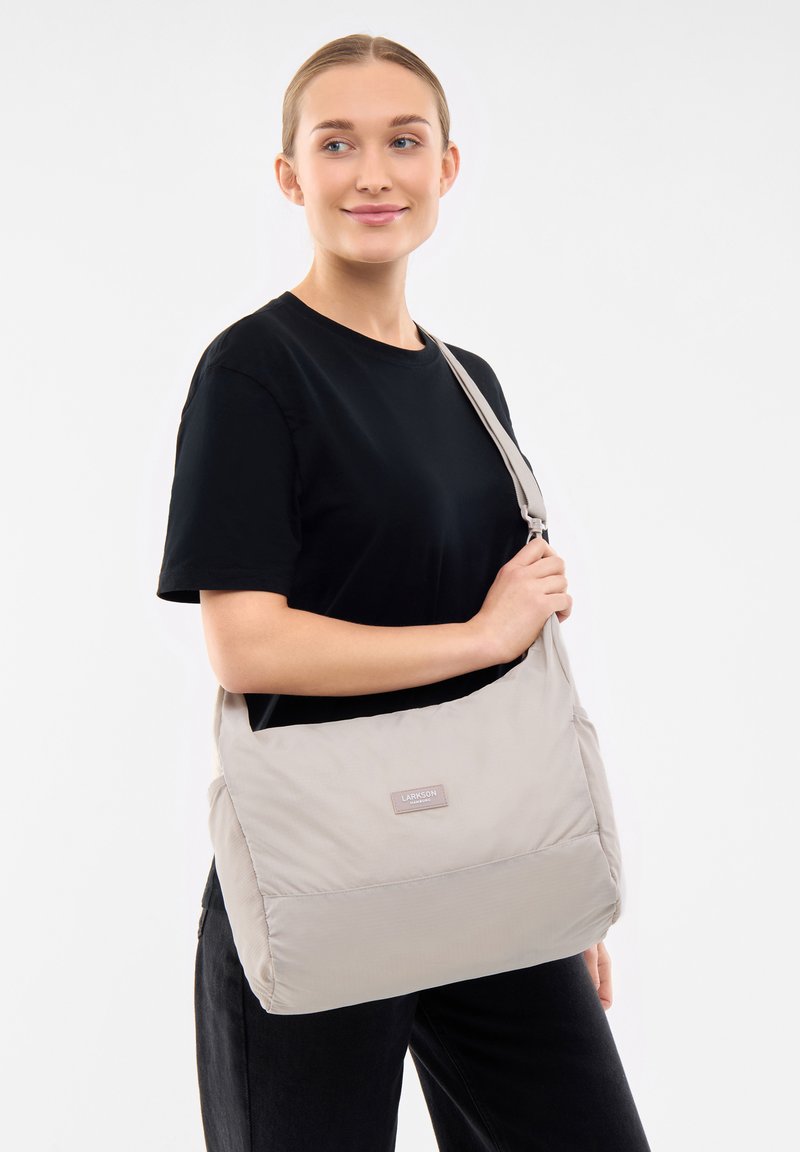 Borsa a spalla in nylon beige con texture liscia, tracolla regolabile e patch logo discreto, caratterizzata da un design spazioso e dai bordi morbidi.