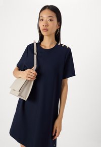 Rochie navy cu mânecă scurtă, guler rotund, cu trei butoane aurii pe umăr, asortată cu o geantă bej deschis. Textură netedă.