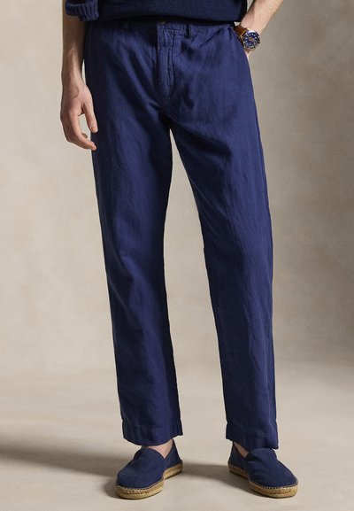 Mężczyzna w granatowych spodniach i pasujących wsuwanych butach, z jedną ręką w kieszeni i zegarkiem na nadgarstku.