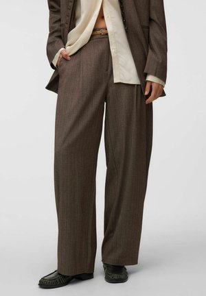 Personne portant un pantalon marron à jambes larges à fines rayures, des chaussures noires, une chemise crème boutonnée rentrée et un blazer marron, debout avec une main dans la poche.