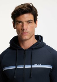 Mörkblå zip-up hoodie med en framlogotyp, två ljusblå horisontella ränder och huva med dragsko. Mjukt tyg och figursydd design.