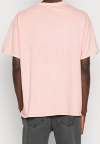 Muž oblečený v jednoduchém světle růžovém tričku s krátkým rukávem a tmavě šedých džínách, zobrazený zezadu na neutrálním pozadí.