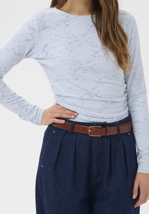 Kvinde iført en lyseblå blomstret blonddetaljeret langærmet top, stoppet ned i højtaljede mørkeblå jeans med et brunt læderbælte.