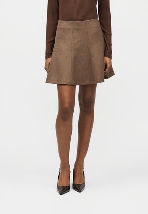 Femme portant une mini-jupe trapèze marron, un haut à manches longues côtelé et des escarpins noirs à bout pointu sur un fond uni.