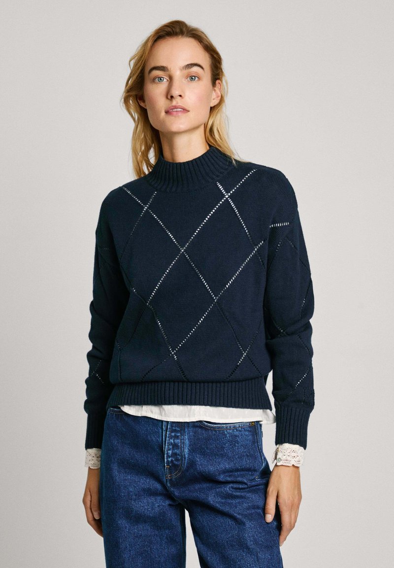 Marineblauwe trui met hoge hals, voorzien van een ruitpatroon en een geribbelde zoom. Draagt over een witte blouse met kraag. Blauwe spijkerbroek zichtbaar.